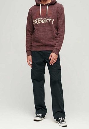 Mężczyzna w bordowej bluzie z kapturem Superdry, czarnych spodniach typu cargo oraz czarnych tenisówkach z białymi sznurowadłami, stojący na jasnym, jednolitym tle.