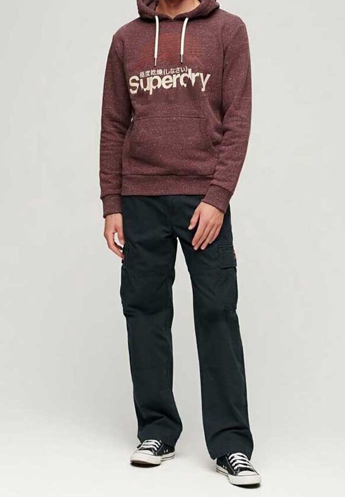 Homme portant un sweat à capuche Superdry couleur bordeaux, un pantalon cargo noir et des baskets noires avec des lacets blancs, debout devant un fond clair uni.