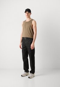 Tan ermeløs tanktop med logo, kombinert med svarte denimbukser med lys søm. Bært med hvite treningssko og en svart caps.