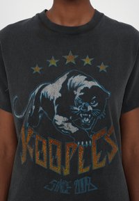 Černé tričko s grafikou syčící panter, nad kterým je pět hvězd, a nápisem "Kooples" s oranžovými a modrými akcenty, nošené osobou.