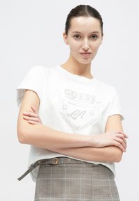Γυναίκα με σκούρα μαλλιά, φορώντας λευκό T-shirt Guess με σχέδιο αστεριού, σταυρωμένα χέρια και γκρι καρό φούστα με ζώνη, μπροστά σε λευκό φόντο.