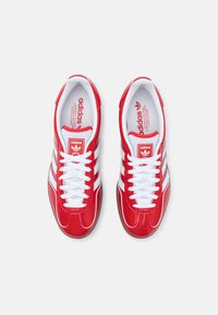 Red athletic shoes with a glossy finish, white mesh tongue, white laces, and three white stripes design on the sides. Red logo on the tongue.