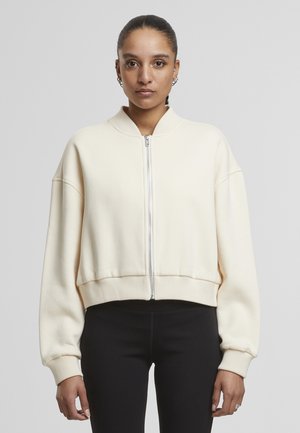 Sweat-shirt court couleur crème avec une fermeture éclair à l'avant, des poignets côtelés et un col montant. Le tissu a une texture douce et lisse.