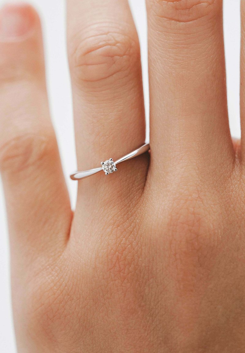Anillo de plata con una banda delgada y un pequeño diamante redondo engastado en un ajuste de seis garras, llevado en una mano de piel clara. Textura de la piel visible.