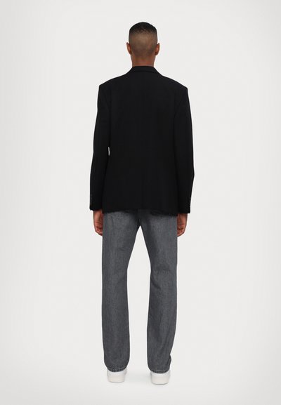 Zwarte op maat gemaakte blazer met een zachte textuur, gecombineerd met grijze rechtgesneden jeans en witte sneakers. Weergave van de achterkant die de pasvorm benadrukt.