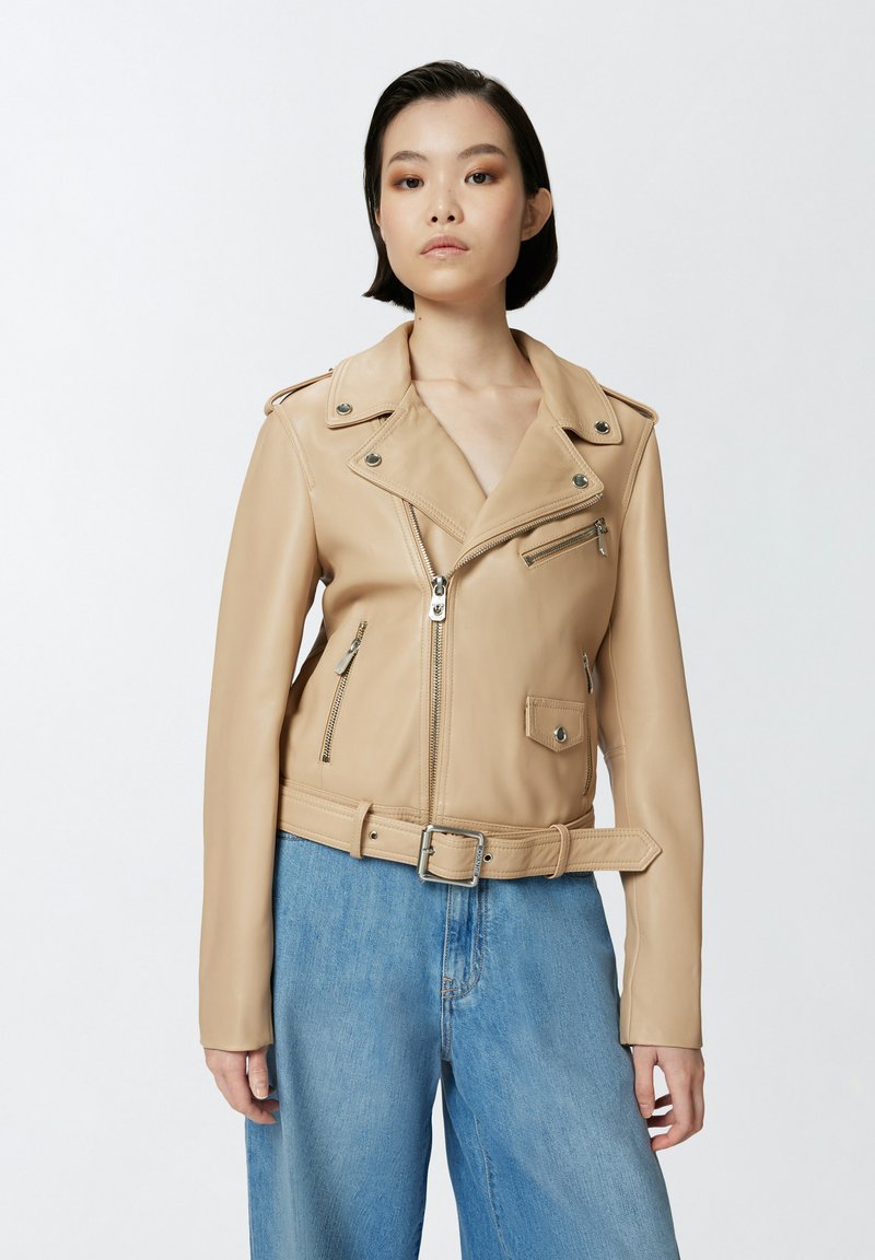 Chaqueta de motorista de cuero marrón claro con cuello de solapa, bolsillos con cremallera y cintura con cinturón. Textura suave y diseño ajustado. Combinada con pantalones de pierna ancha azules.