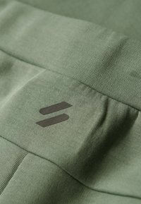 Tessuto verde con una superficie liscia, caratterizzato da un logo nero prominente al centro. Il materiale presenta una leggera elasticità e morbidezza.