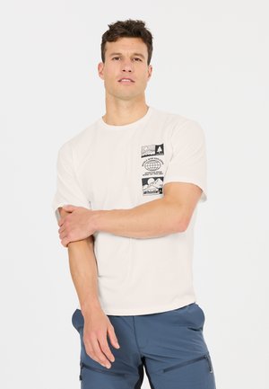 Mann trägt weißes T-Shirt mit Außenmotiven und blauen, mit Reißverschluss versehene Hosen, steht vor weißem Hintergrund.