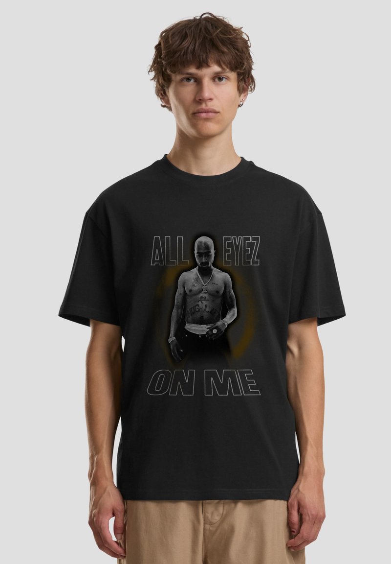 Junger Mann trägt ein schwarzes T-Shirt und zeigt ein Schwarz-Weiß-Foto eines oberkörperfreien Mannes mit Tätowierungen und dem Text „ALL EYEZ ON ME.“