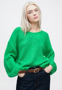 Πράσινο πλεκτό πουλόβερ με χαλαρή εφαρμογή, στρογγυλή λαιμόκοψη και μακριά μανίκια, συνδυασμένο με σκούρα τζιν και καφέ ζώνη.