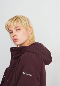 Junge Person mit kurzem blonden Haar, die eine dunkellila Columbia-Jacke mit Kapuze trägt und leicht seitlich vor einem schlichten, hellen Hintergrund steht.