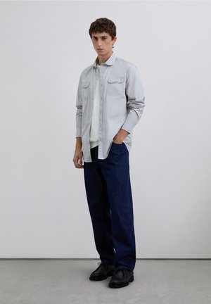 Lichtgrijs overhemd met knopen en twee borstzakken over een wit kledingstuk, gecombineerd met donkerblauwe spijkerbroek en zwarte schoenen.