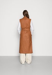 Chaleco largo de cuero marrón claro con cintura ceñida y sin mangas, combinado con una camisa blanca y pantalones beige de corte holgado.
