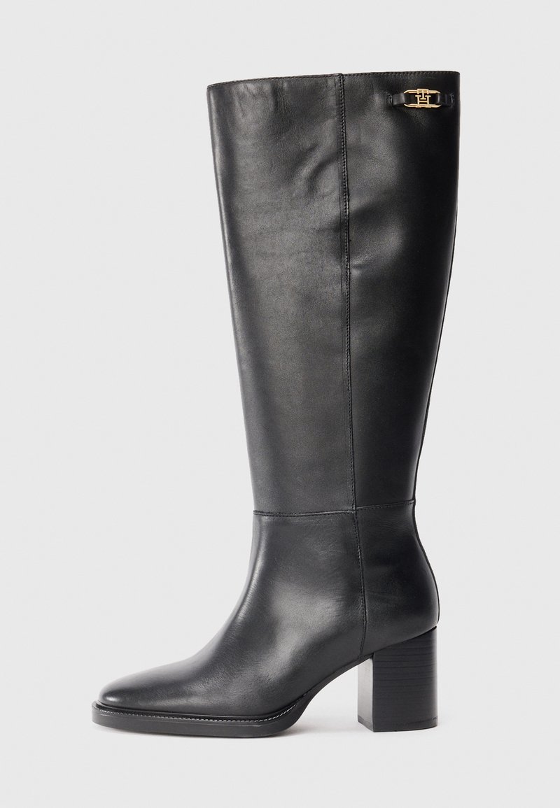 Botte en cuir noir, arrivant au genou, avec un talon bloc, un bout pointu et un accent en métal doré près du haut. Texture lisse avec un minimum de coutures.