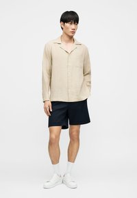 Homme portant une chemise beige à manches longues boutonnée, un short bleu marine, des chaussettes blanches et des baskets blanches, debout devant un fond blanc uni.