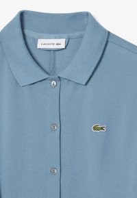 Polo shirt blu chiaro con tessuto texture, dotato di colletto a costine, due bottoni grigi e un piccolo logo di coccodrillo verde sul petto.