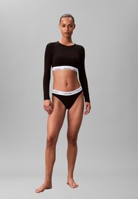 Black long-sleeve crop top with white Calvin Klein logo band, paired with black bikini bottoms featuring a matching logo waistline.