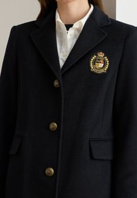 Μπλε ναυτικό μάλλινο σακάκι με χρυσά κουμπιά, γιακά με πέτο και σήμα με έμβλημα στο αριστερό στήθος. Διακρίνονται δύο πλαϊνές τσέπες.