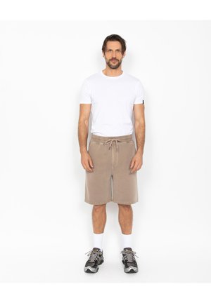 Homme debout portant un t-shirt blanc, un short beige à cordon, des chaussettes blanches et des baskets grises sur un fond blanc uni.