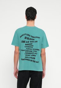 Τιρκουάζ βαμβακερό μπλουζάκι με κοντά μανίκια και στρογγυλή λαιμόκοψη, με έντονο μαύρο κείμενο σε ποικίλη διάταξη στην πλάτη, χωρίς επιπλέον γραφικά.