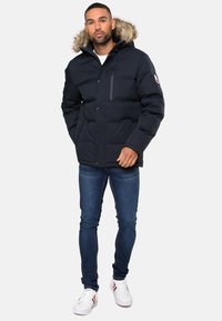 Marineblaues Puffer-Jackett mit Kapuze mit Fellbesatz, durchgehender Knopfleiste und einer Seitentasche; kombiniert mit blauen Jeans und weißen Sneakers.