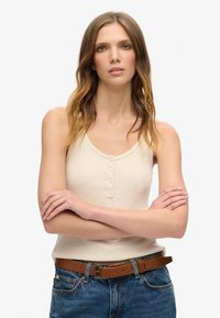 Lys beige ribbet tanktop med rund halsudskæring og knapdetaljer foran, parret med højtaljede blå denim jeans og et brunt bælte.