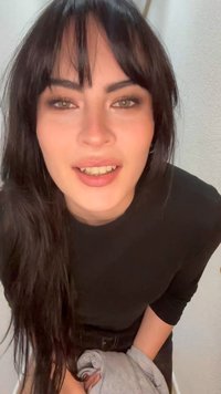 Jeune femme aux longs cheveux foncés avec une frange penchée en avant, souriante, portant un haut noir et tenant une veste grise devant un mur clair.