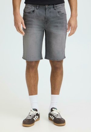 Mężczyzna ubrany w wyblakłe szare szorty dżinsowe, białe skarpety za kostkę oraz czarne buty sportowe Adidas z białymi paskami, stojący na tle o neutralnym kolorze.