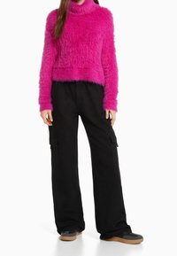 Flauschiger rosa Rollkragenpullover mit langen Ärmeln, kombiniert mit schwarzen Cargo-Hosen mit Taschen und einem lockeren Schnitt. Schuhe mit brauner Sohle.