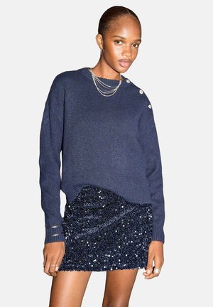 Pull en tricot bleu marine avec des accents de boutons à l'épaule, associé à une mini-jupe bleu marine à sequins, présentant une finition texturée.