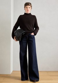 Maglione a collo alto ribbed marrone scuro, jeans a gamba larga in denim scuro. Il modello tiene una pochette in pelle nera, in piedi su un pavimento in legno chiaro.