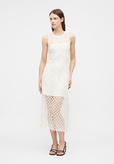 Femme portant une robe midi sans manches en dentelle blanche avec une jupe inférieure transparente et des sandales à talons noires à lanières, debout devant un fond uni.