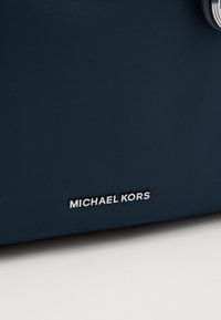 Sac en matériau texturé bleu marine avec un logo métallique argent "MICHAEL KORS", présentant un design épuré et des accents matériels minimalistes.