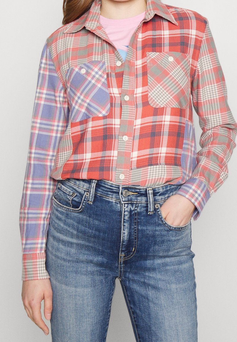 Femme portant une chemise à carreaux multicolore avec des tons bleus et rouges et un jean bleu taille haute, une main dans la poche.