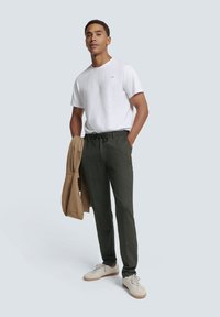 White t-shirt, drawstring dark green pants, beige jacket over the arm, and light sneakers in a neutral setting.