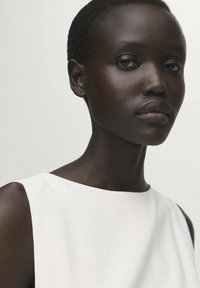 Indumento senza maniche bianco con scollatura rotonda. Il tessuto liscio e le linee pulite mettono in evidenza un design minimalista. Carnagione scura e capelli corti visibili.