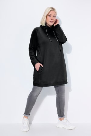 Frau mit blondem Haar, die ein schwarzes langes Hoodie-Kleid, graue Skinny-Jeans und weiße Sneaker trägt, steht mit einer Hand in der Tasche.