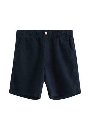 Teenagedreng stående med hænderne i lommerne, iført navyblå kortærmet polo, mørke shorts, hvide sokker med striber og sorte sneakers på hvid baggrund.