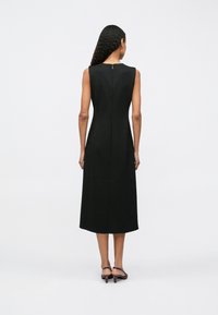 Žena s dlouhými kudrnatými vlasy, oblečená v bezramínkových černých midi šatech a hnědých sandálech na nízkém podpatku, stojící zády k bílé zdi.