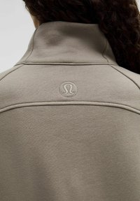 Cremefarvet sweatshirt med høj krave, der har stiksyninger og et lille logo på ryggen. Blød stoftekstur.