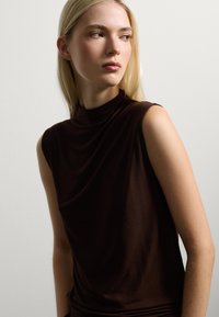 Haut sans manches marron avec un col haut, fabriqué en tissu lisse, présentant des drapés doux et une texture subtile, ajusté au niveau des épaules.