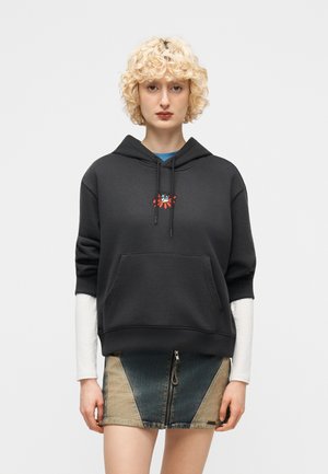 Oseba z blond kodrastimi lasmi, oblečena v črno majico s kapuco z majhno grafiko mačke in cvetja, pod njo bele dolge rokave, ter dvobarvno kratko denim krilo.