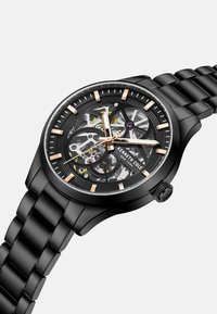 Orologio scheletro nero con bracciale in metallo nero, dettagli in tono oro, quadrante trasparente che rivela i meccanismi interni e una lunetta testurizzata.
