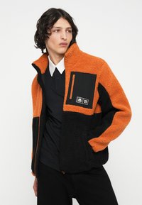 Chaqueta de forro polar con colores naranja y negro, bolsillo con cremallera en el pecho y tejido texturizado con diseño de paneles en contraste.