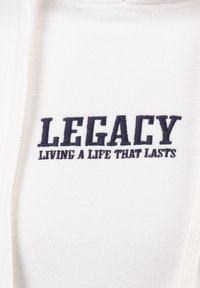 Witte hoodie met donkerblauwe borduurring. De tekst luidt "LEGACY" boven "LIVING A LIFE THAT LASTS." Zachte stof met een gladde textuur.