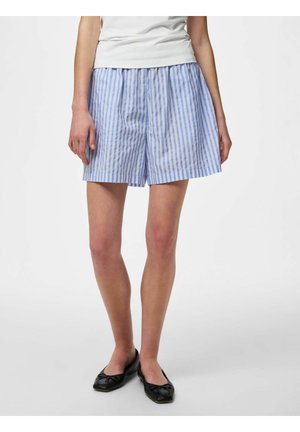 Kvinne som har på seg løstsittende blå og hvite vertikalt stripede shorts, hvit topp og svarte ballettsko, stående mot en ensfarget bakgrunn.