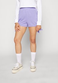 Kvinna i lila Adidas-shorts, vit långärmad tröja, vita strumpor och kraftiga off-white sneakers står framför en enfärgad bakgrund.