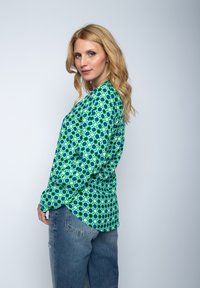 Długi rękaw koszula z błyszczącego zielonego materiału z okrągłym wzorem w niebieskim i białym kolorze. Ma luźny krój i zaokrąglony dół.