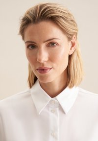 Weißes Hemd mit Knopfleiste und spitzen Kragen, aus glattem Stoff gefertigt. Hat ein klares Design mit einem subtilen Glanz und kleinen Knopfdetails.