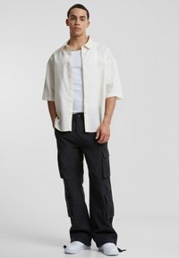 Kortärmad, ljusbeige linnen skjorta med knappar, i kombination med svarta cargobyxor som har flera fickor och vita sneakers.